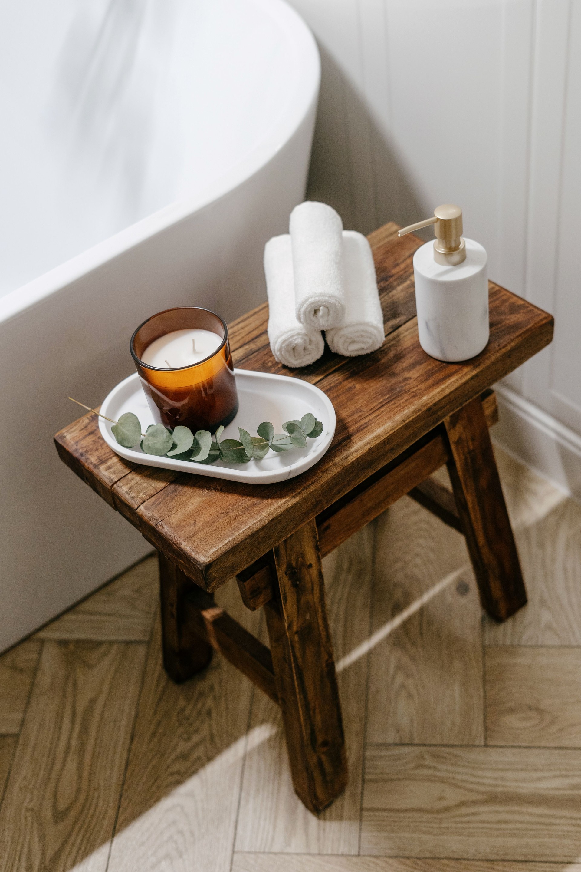 Soap dispenser bottle, towels and scented candle on wooden bench near white tub